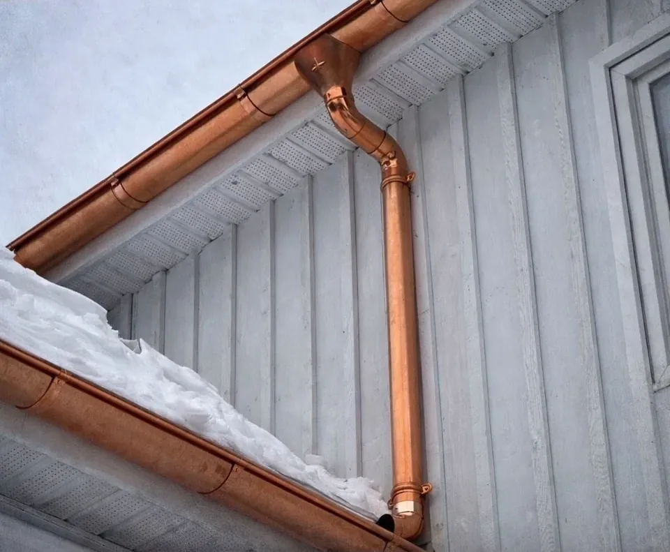 A copper seamless gutter and downspout running along the corner of a white board-sided home, snow drifted along the roofline